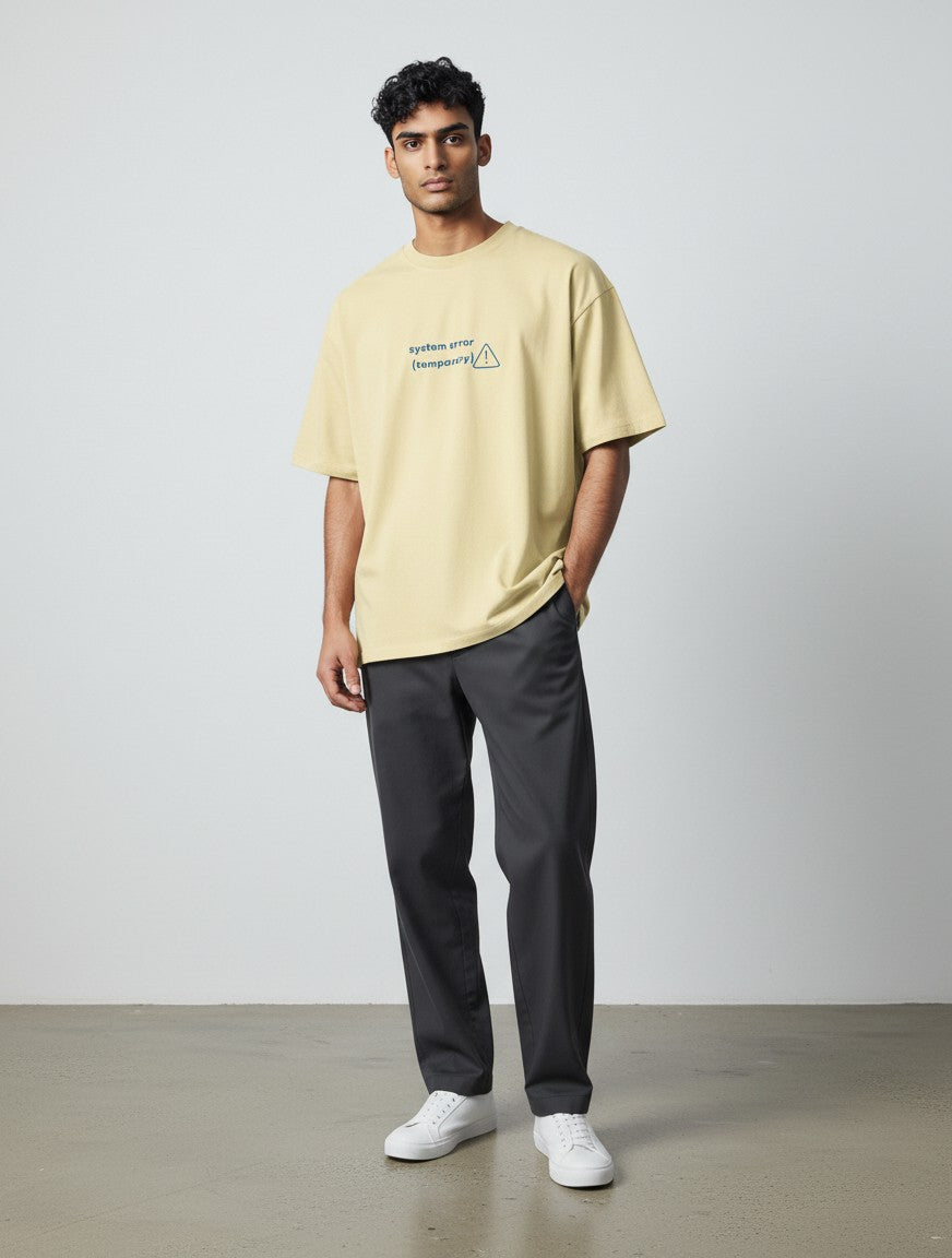 A man wearing a beige mi-DONE oversized cotton t-shirt with a System Error graphic, paired with dark grey trousers.