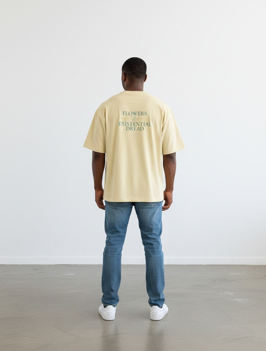A man wearing a beige mi-DONE oversized cotton t-shirt with Flowers and Existential Dread text on the back.