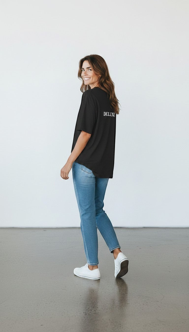 A woman wearing a black mi-DONE oversized cotton t-shirt with white text on the back, paired with blue jeans and sneakers.
