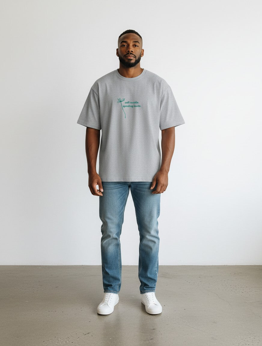 A man wearing a grey mi-DONE oversized cotton t-shirt with a small green graphic print, paired with blue jeans.