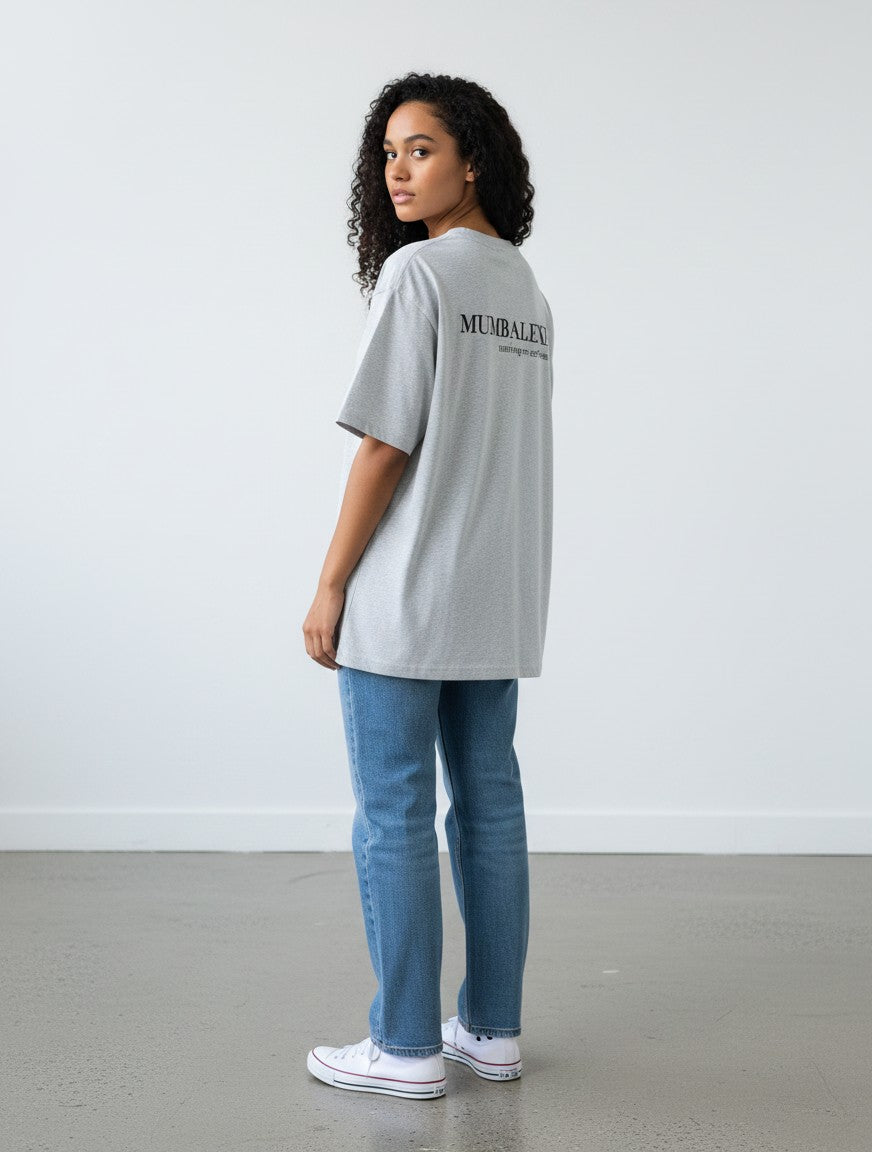 Model wearing a grey mi-DONE oversized cotton t-shirt with Mumbai print, paired with blue jeans and white sneakers.