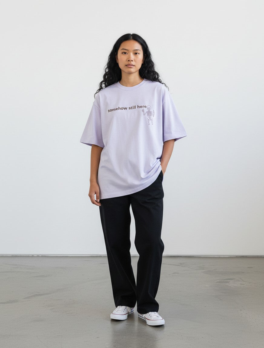 mi-DONE oversized lavender cotton t-shirt with skeleton graphic and somehow still here text worn by a woman
