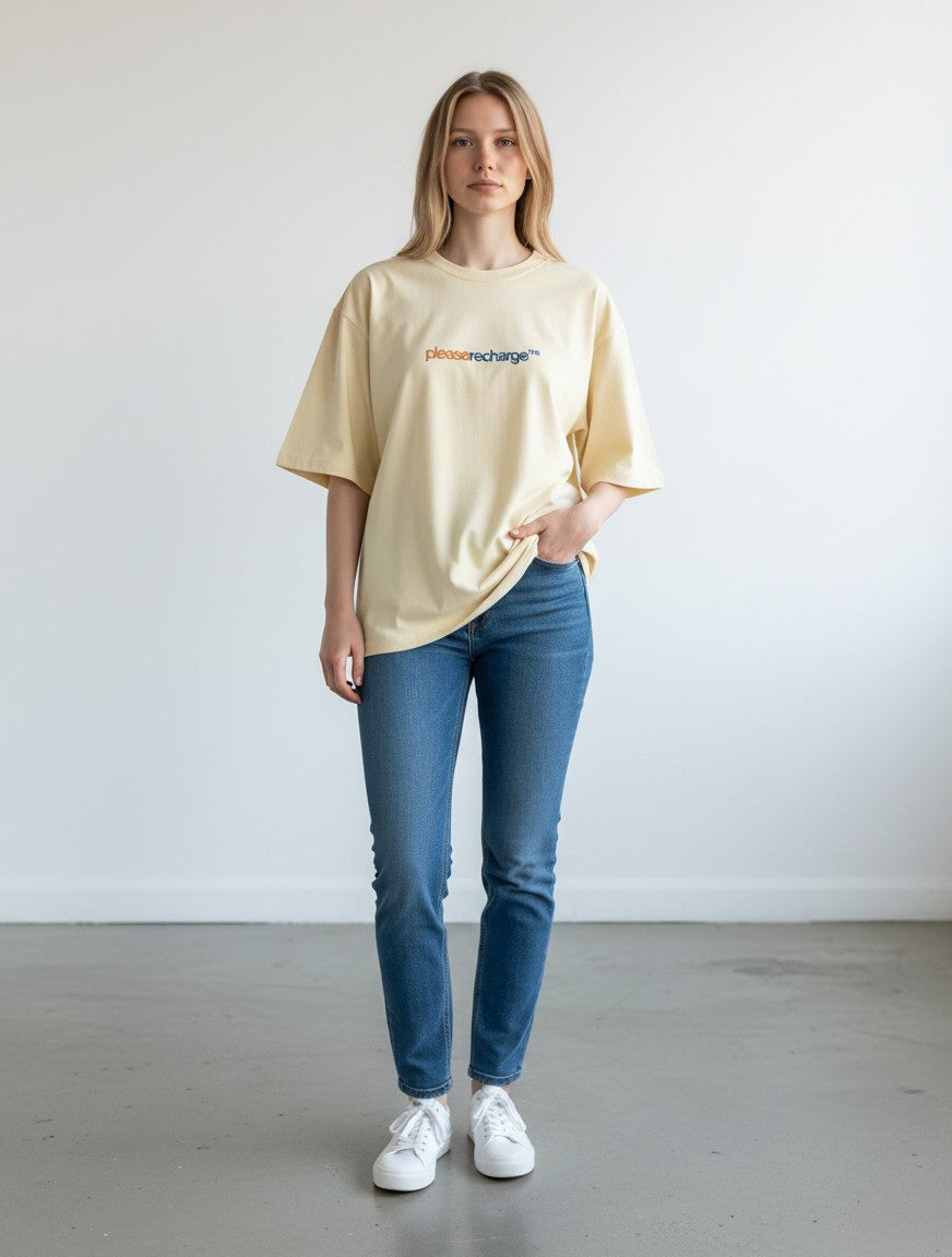 A woman wearing a mi-DONE oversized cream cotton t-shirt with please recharge graphic, paired with blue jeans and sneakers.