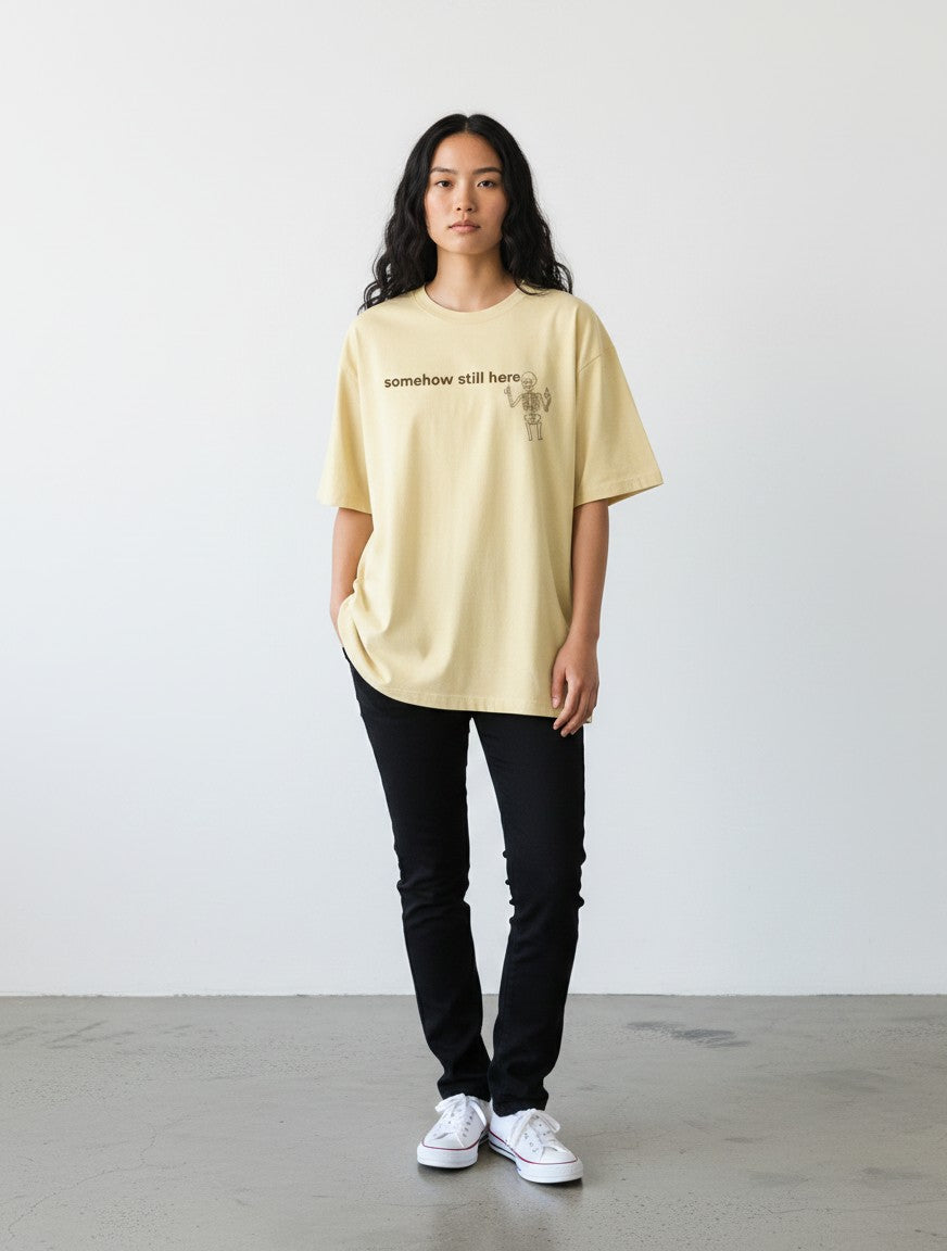Model wearing a mi-DONE oversized yellow cotton t-shirt with a skeleton graphic and somehow still here text.